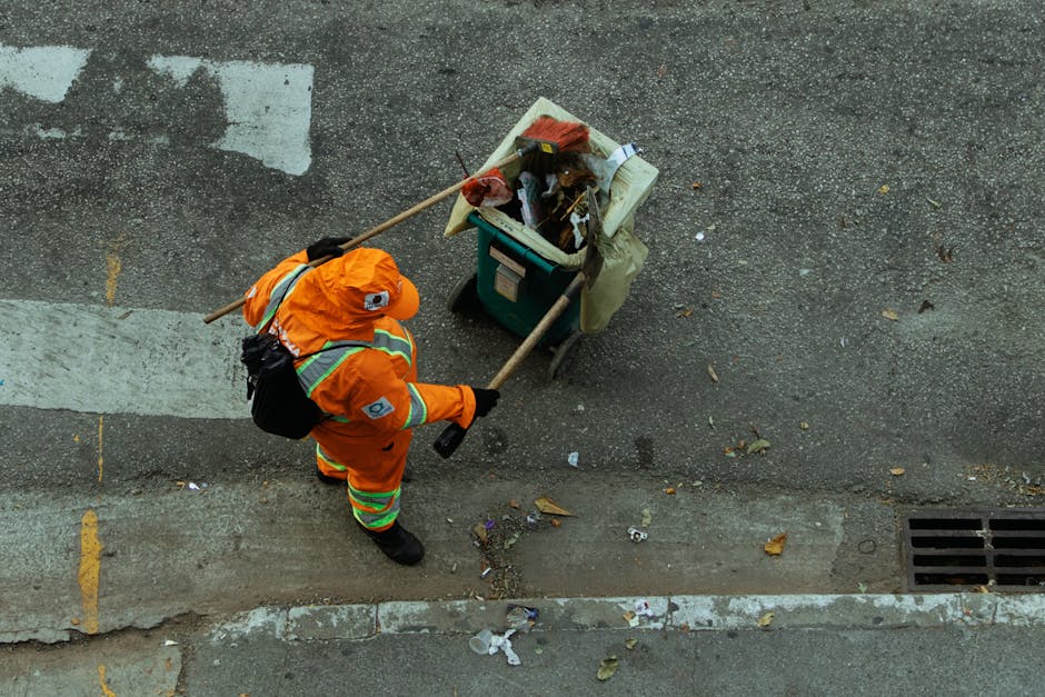 A municipal street cleaner wearing an orange reflective safety jacket and matching cap is seen from above, sweeping up scattered litter, including paper, leaves, and a plastic container, on the sidewa