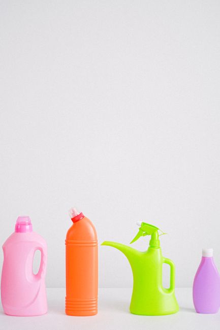 A collection of colorful plastic chemical containers and jugs, including blue, yellow, red, and black bottles, arranged outdoors on the ground, likely for waste disposal or cleaning supplies. The cont