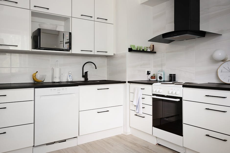 A modern kitchen featuring white flat-panel cabinets with black handles, a black countertop, and a black sink with a curved faucet. The backsplash is made of large, light-colored tiles, and the room is well-lit with natural light. Visible objects include a banana in a fruit bowl, a roll of paper towels, a soap dispenser, a microwave oven positioned above the countertop, and some small kitchen appliances such as a radio and a toaster. To the right, there is a white freestanding oven with black glass doors, a black range hood above it, and a round wall clock on the wall. A white towel hangs from the oven handle, and the floor appears to be light wood or laminate. The surfaces are clean, and the overall appearance suggests recent surface cleaning and maintenance executed by Oven Cleaning Islington, aligning with the surface cleaning and hygiene standards described in the Angel N1 guide for flats and kitchens.