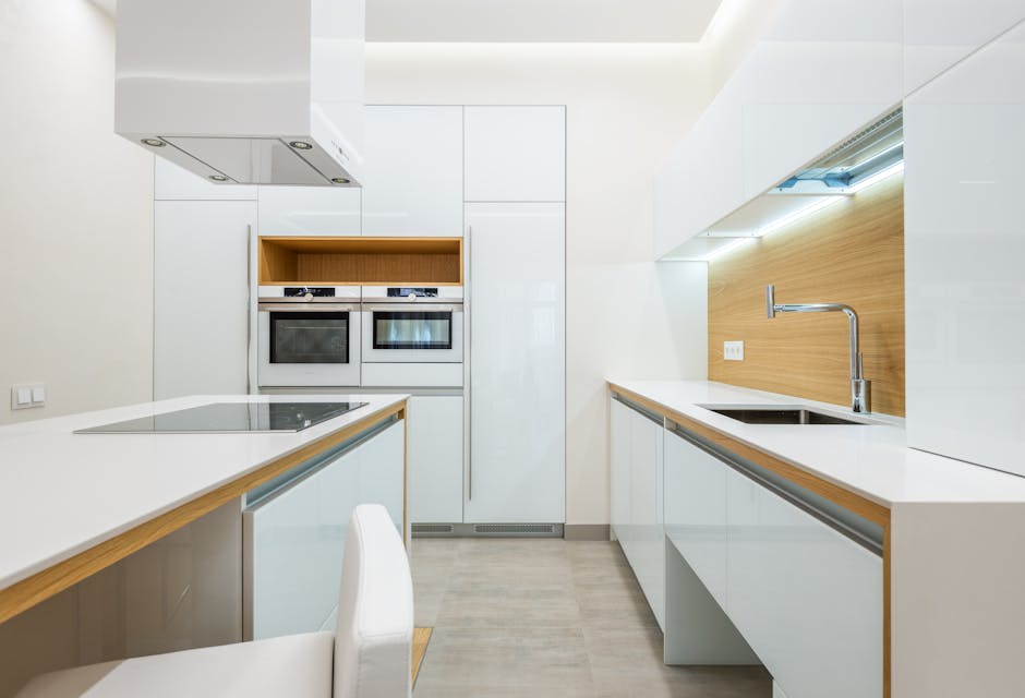 A modern kitchen with white glossy countertops and cabinets, featuring stainless steel appliances including an oven and microwave built into a tall white pantry. The countertops appear clean and polished, with a smooth surface reflecting light from the ceiling. On the right side, a stainless steel sink with a sleek faucet is integrated into the counter, set against a light wooden backsplash that adds warmth to the space. The lighting is bright and even, highlighting the pristine condition of all surfaces. The flooring is a light-colored wood or vinyl, complementing the overall clean appearance of the kitchen. Visible in the scene are minimal decorative elements, emphasizing the hygienic and well-maintained environment, consistent with the professional cleaning services provided by Oven Cleaning Islington. This setup exemplifies expert surface cleaning and sanitisation, ideal for domestic and commercial kitchens as described in the oven cleaning Angel N1 guide for flats and kitchens.