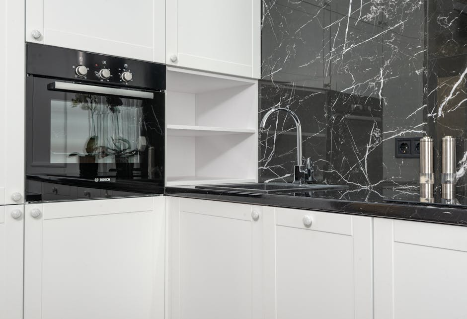 A modern kitchen featuring white cabinetry and a black marble backsplash with white veining. The black glass-ceramic cooktop is integrated into the black marble countertop, which appears clean and pol